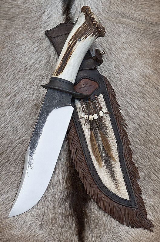 a large hunting or survival knife rests on a bed of gray fur. the knife has a polished silver blade and a handle made from a deer antler, with brown streaks and a crown of knobs at the top. a black guard separates the blade from the handle. the knife is partially sheathed in a decorative leather sheath that is adorned with a panel of contrasting light fur, fringe, and strings of beads.