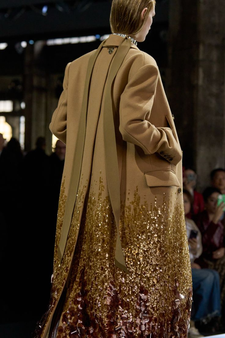 a woman wearing a long brown coat with a gold sequin hem. the coat is open in the back, revealing the sequin hem. the woman is wearing a black choker and is walking on a runway.