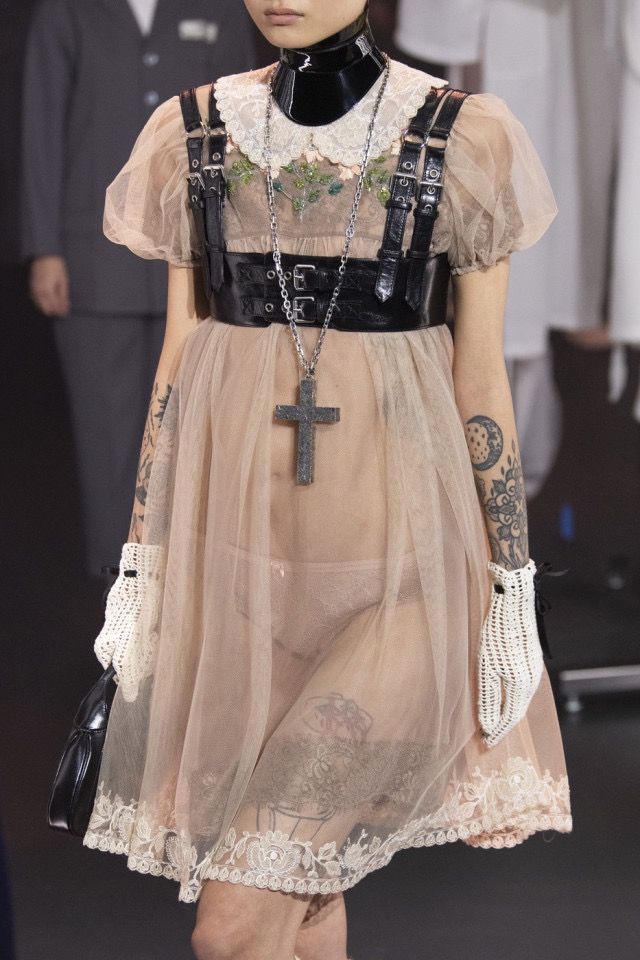a model is walking on a runway during a fashion show. she is wearing a short, light pink transparent dress with lace trim. the dress has a white lace collar and is adorned with a black leather harness. she has numerous tattoos on her arms and legs. she is also wearing a black choker, a long silver necklace with a large cross pendant, and white crochet gloves. she is carrying a small black purse. the background is blurred, but other people and clothing can be seen.