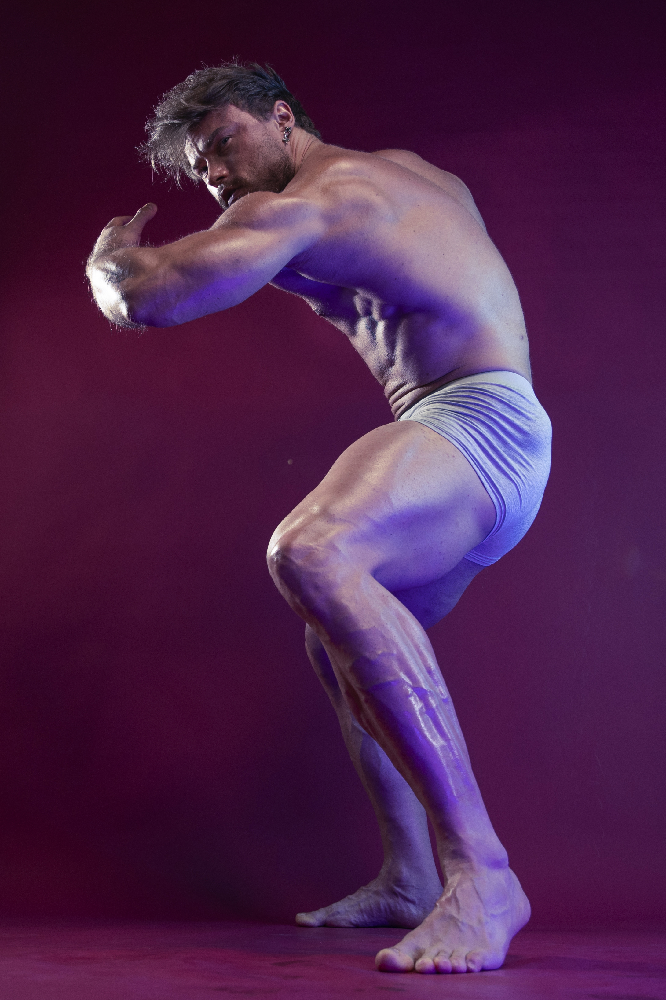 a muscular man with a welldefined physique is posing in a studio setting. he is shirtless, wearing only underwear, and flexing his muscles. the background is a solid purple color. he is standing with a bent posture, with his legs spread apart and arms bent at the elbows, showing off his biceps. the lighting is focused and highlights the contours of his body.