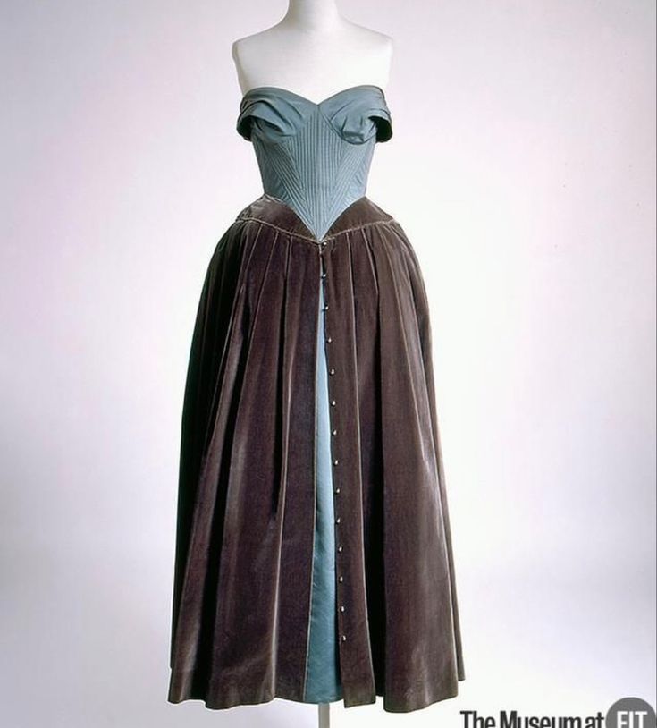 a strapless teal dress with a brown velvet overskirt on a mannequin is displayed in front of a white background. the bodice of the dress is teal and features a sweetheart neckline. the brown velvet overskirt has buttons down the front. the dress is on display at the museum at fit.