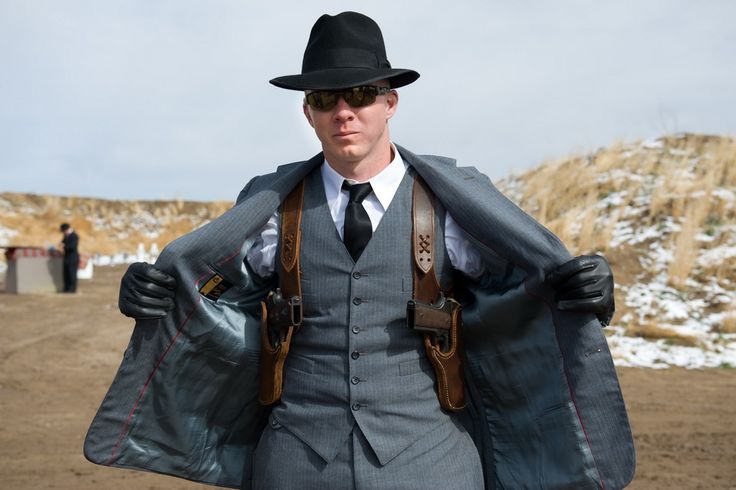a man in a gray suit and black fedora is standing outdoors at a target range. he is wearing sunglasses and leather gloves. he is opening his jacket to reveal two handguns in shoulder holsters. another man is standing at a target in the background.
