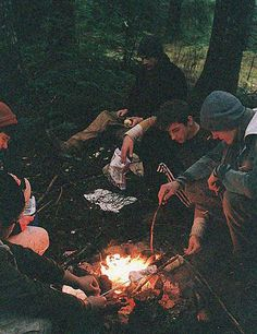 the image depicts a group of young adults gathered around a crackling campfire in a dark forest setting.  they appear to be camping or backpacking, possibly roasting marshmallows or other food over the flames. the atmosphere is intimate and cozy, with the warm firelight contrasting against the surrounding darkness of the night. the friends are dressed in casual, layered clothing suitable for cooler weather. the scene conveys a sense of camaraderie and enjoyment of the outdoors.