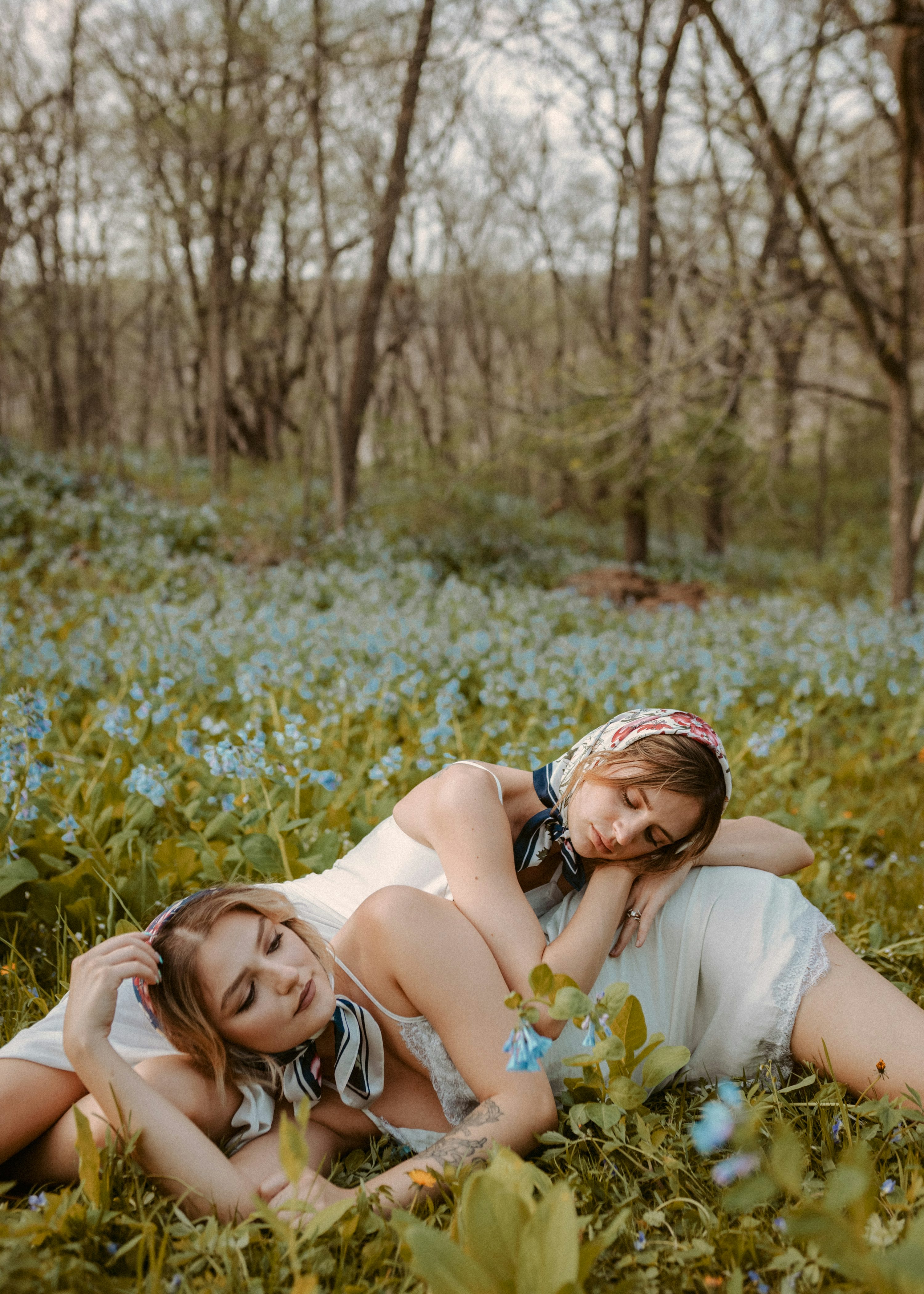 two women are lying down in a field of blue flowers, embracing each other. they are wearing white dresses and floral headscarves. the setting is a peaceful forest with trees and sunlight. the overall mood is romantic and serene.
