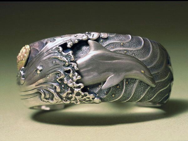 a closeup shot features a silver bracelet with a nautical theme. the bracelet showcases a dolphin leaping through a wave. the wave is intricately detailed with textured silver to resemble splashing water. the background of the bracelet has a curved pattern. there are small gold accents. the bracelet appears to be handmade or artisancrafted.