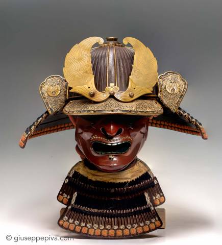 the image showcases a meticulously crafted antique japanese samurai helmet and face mask displayed on a stand.  the helmet, adorned with ornate golden crests, sits atop a menacing red and black lacquered face mask. the mask features exaggerated features, including a fierce expression with a grimacing mouth. below the mask, a partial suit of samurai armor is visible, hinting at the protective gear worn by these legendary warriors. the image is captured against a neutral gray background, drawing the viewer's attention to the intricate details and craftsmanship of the artifacts.