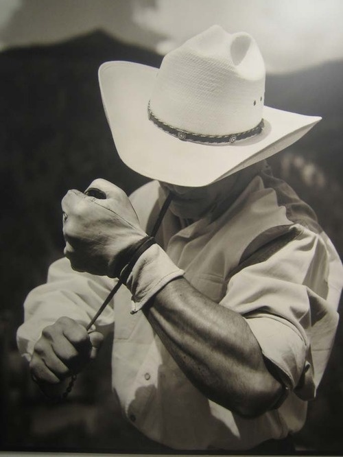 the image is a striking black and white closeup photograph of a cowboy. he's wearing a widebrimmed white cowboy hat, lightcolored longsleeved shirt, and gloves.  his muscular arm is prominently featured as he appears to be in the midst of roping or working with a lasso (a thin rope or cord is partially visible). the background is blurred, suggesting an outdoor setting, likely on a ranch or in a rodeo environment. the overall mood is one of strength, skill, and the traditional imagery associated with the american west. the lighting and contrast in the photograph emphasize the texture of the cowboy's clothing and skin and the strength in his arm.