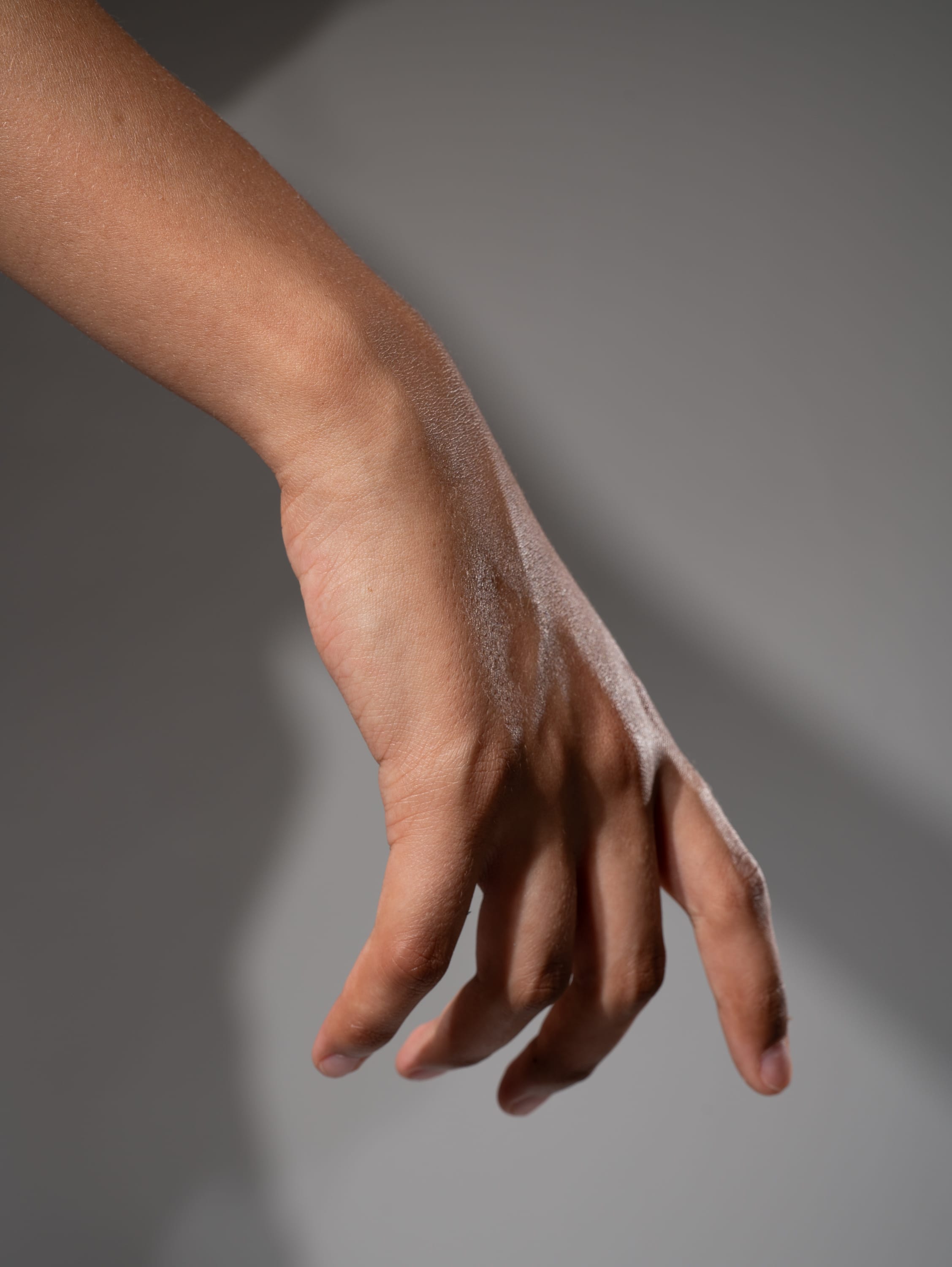 a closeup image of a hand reaching downwards against a gray background. the hand is positioned with the fingers slightly curled inward, creating a sense of anticipation or grasping. the lighting is soft, highlighting the contours and texture of the skin.