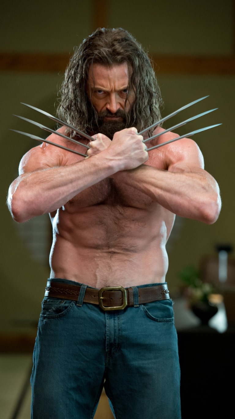 this is a full shot of hugh jackman as wolverine, his arms crossed with his adamantium claws extended. he is shirtless, wearing jeans and a brown belt. he has a beard and long, unkempt hair. his expression is fierce and determined. the background is blurred, suggesting an interior setting.