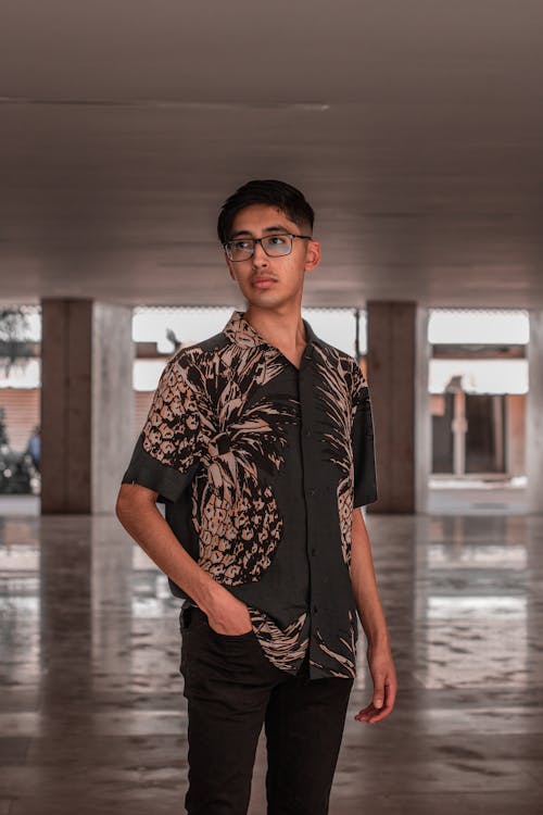 a young man stands in a spacious hallway, wearing a dark green buttondown shirt with a pineapple print and black pants. he wears glasses and has a thoughtful expression, looking slightly to his right.  the hallway is modern and has a reflective floor. the overall mood is casual and stylish.