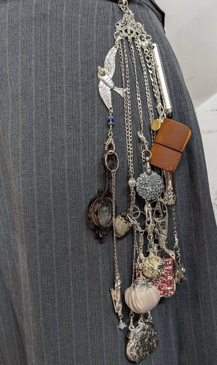 a closeup shot of a grey pinstripe skirt with an ornate silver charm collection hanging from the waistband. the charm collection includes a silver bird, a small leather notebook, a metal cage, a coin purse, a heart, and a scissor charm, all hanging from various silver chains.