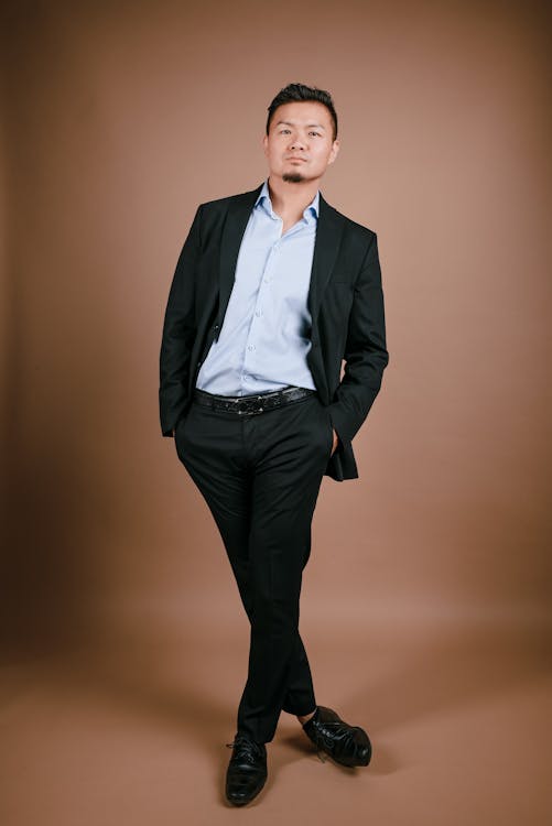 a fulllength portrait of an asian man wearing a black suit, light blue dress shirt, and black dress shoes against a brown background. he is standing with his legs crossed, and his hands are in his pockets. he is looking directly at the camera.
