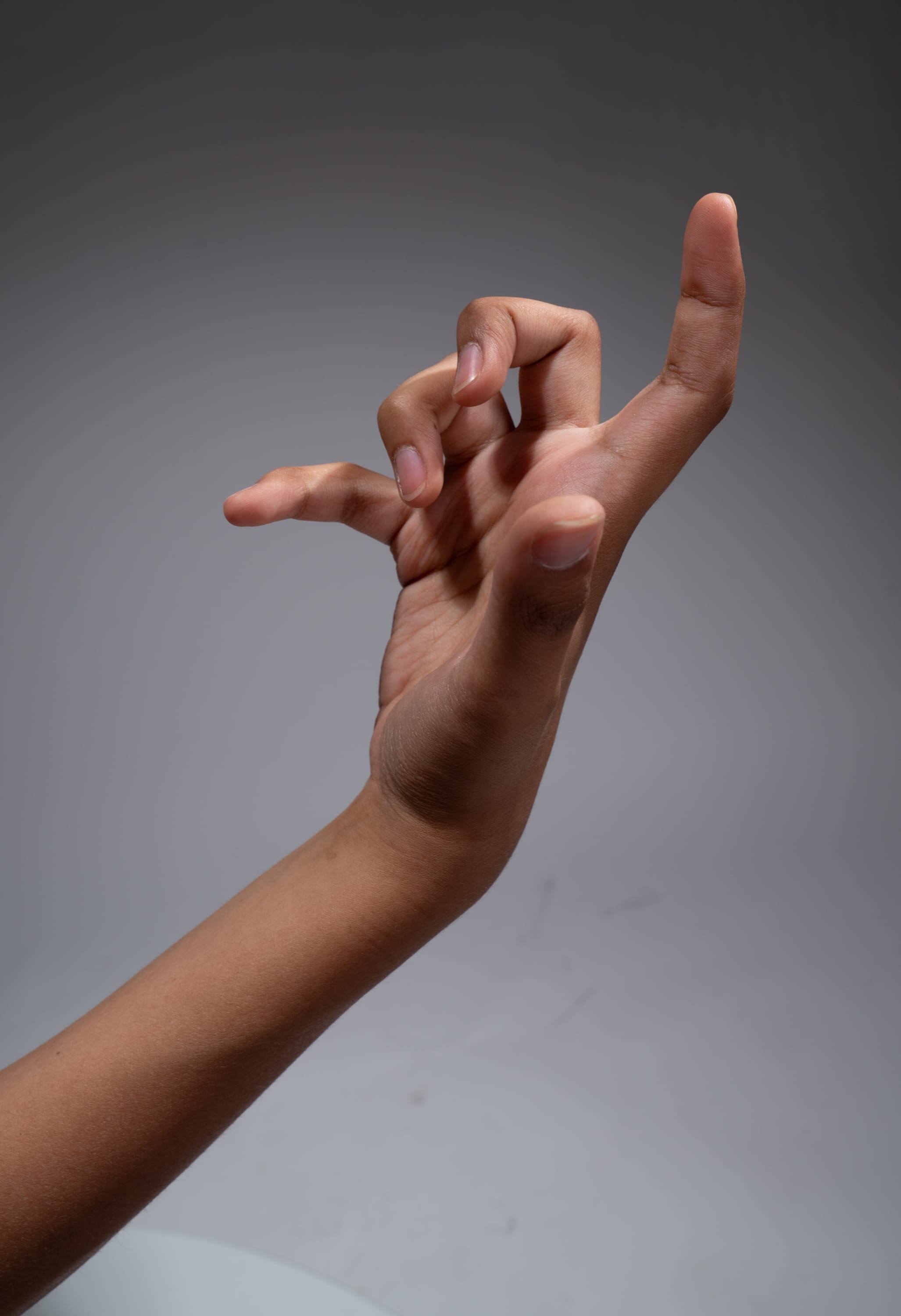 a closeup shot of a hand with brown skin, extended upwards against a light grey background. the hand is posed with the fingers curled in a slightly unusual gesture. the lighting is soft and diffused, highlighting the contours of the hand and arm.