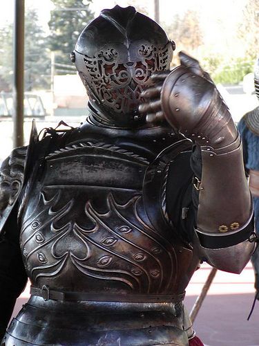 a man dressed in a full suit of ornate, medievalstyle armor. the armor is primarily silver with intricate designs. he stands in an outdoor setting, with a sunny sky visible in the background. he is wearing a helmet with a visor that obscures his face, and he is making a gesture with his right hand.