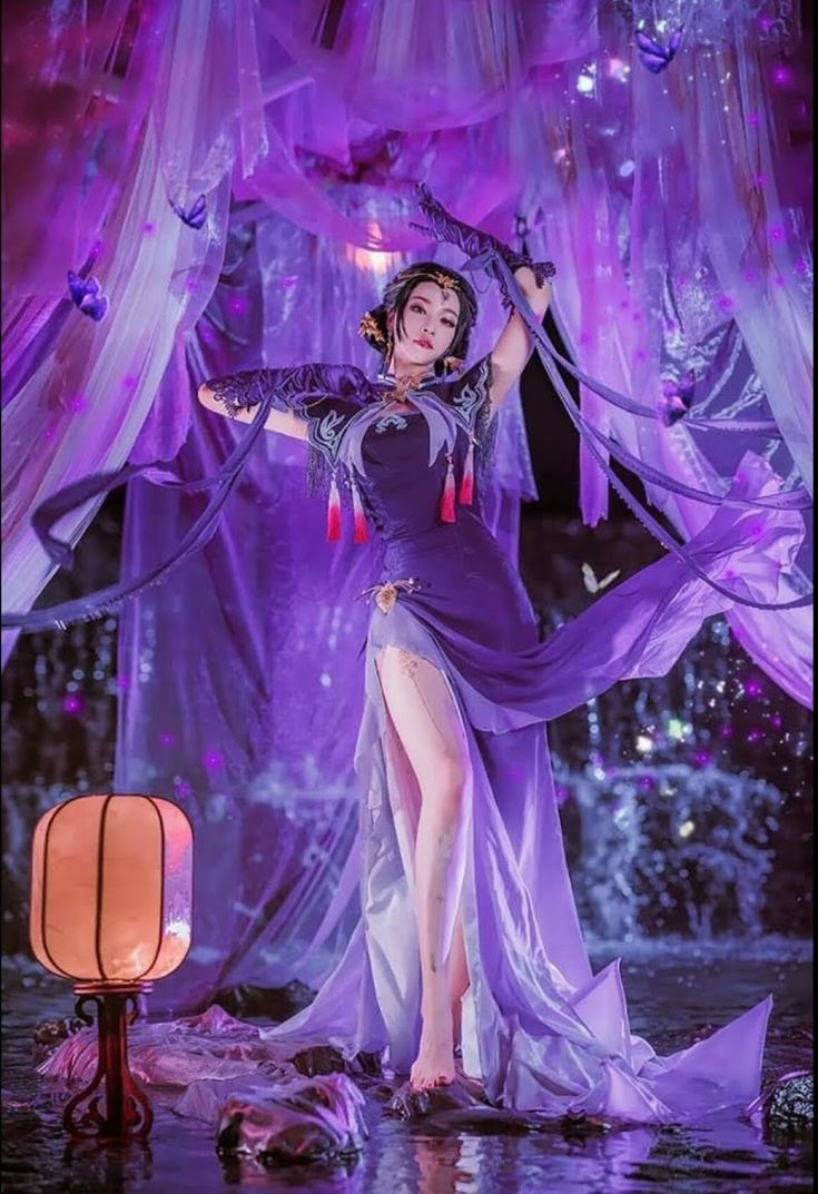 a full shot of a woman in cosplay, dressed in a long, flowing purple dress with a high slit, standing in water. she wears a headdress and gloves, and the dress features tassels and intricate detailing. she is surrounded by fabric drapes and what appears to be butterflies, creating a mystical, magical atmosphere. a lantern is positioned to her left, adding to the stage lighting. the background resembles a fairyland scene.