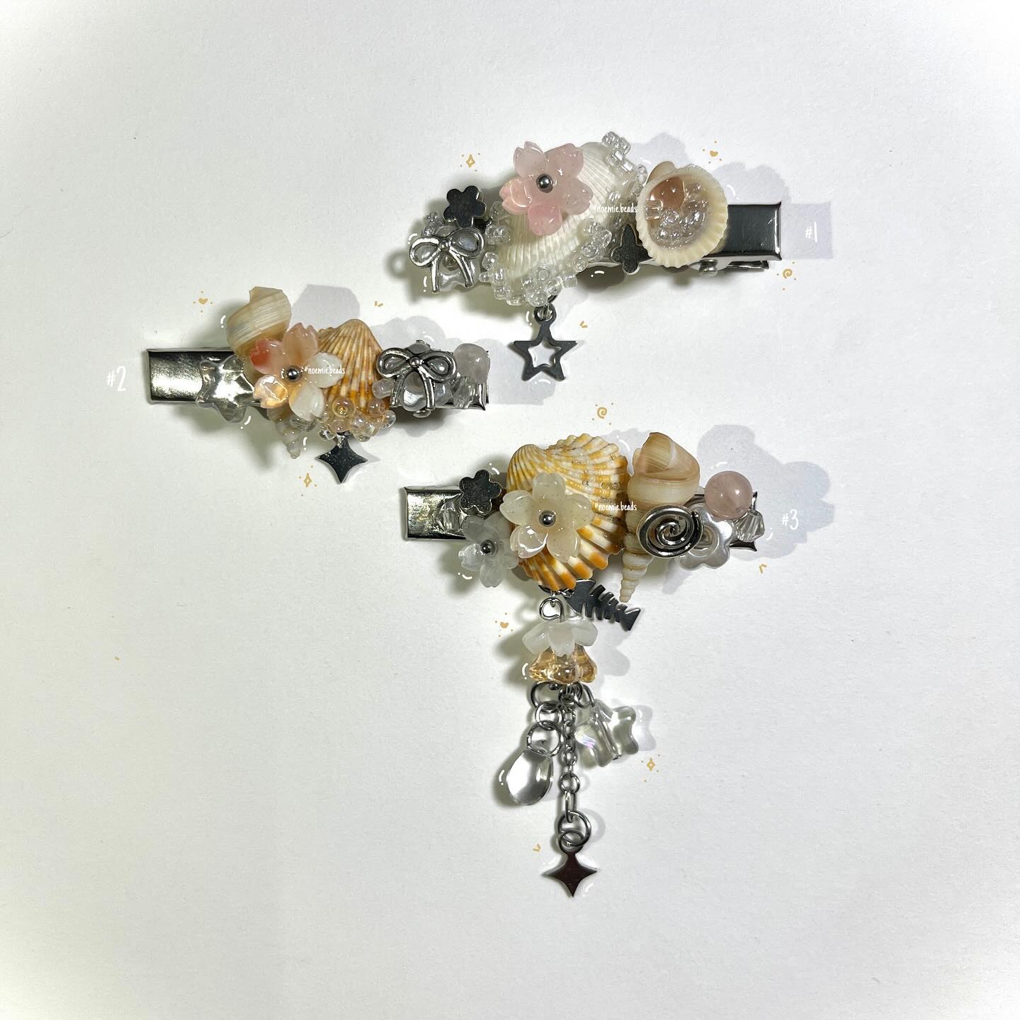 the image showcases three decorative hair clips arranged on a white background. each clip features seashells, flower embellishments, and star charms, along with various beads and silver accents. the clips appear to be handcrafted and are designed to be ornate hair accessories. the composition highlights the intricate details of each clip against the plain backdrop.