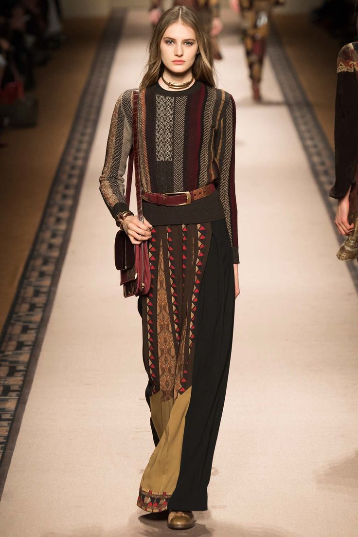a full shot of a female model walking down a runway, showcasing a bohemianstyle outfit. the outfit consists of a patterned sweater with vertical stripes of various colors, a dark skirt, and a long, loosefitting pair of pants with gold and brown patterned fabric. the model is accessorized with a maroon leather crossbody bag, a brown leather belt, and gold choker necklace. her pose and expression suggest confidence and poise. the runway is beige, and the background shows an outoffocus audience and other models.