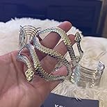 a closeup shot shows a person's hand holding a luxurious, twotoned (gold and silver) snakeshaped bracelet. the bracelet is encrusted with what appears to be diamonds or cubic zirconia, giving it a very sparkly, "icedout" look. the bracelet is intricately designed, with multiple intertwined snakelike bands.  the background is blurry but shows a fluffy white surface and a dark object that seems to be a box or display, potentially indicating a highend jewelry setting.  the overall impression is one of opulence and highend fashion.
