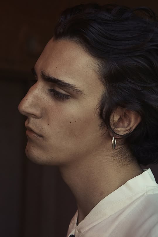 a side portrait of a man with dark hair wearing a white shirt and a hoop earring.