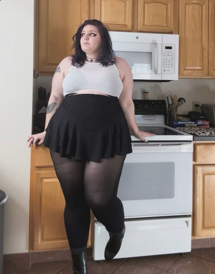 a full figured woman is standing in a kitchen. she is wearing a white crop top, black skirt, tights, and black boots. she is standing in front of a stove and microwave. there are cabinets behind her. she is looking off to the side.