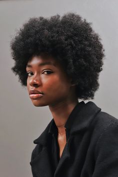 a closeup portrait of a young black woman with a large, voluminous afro. she's wearing a black collared coat, and her expression is serious and confident. the background is a plain, muted gray. the image emphasizes her natural beauty and strong features.  the overall feel is elegant and sophisticated.