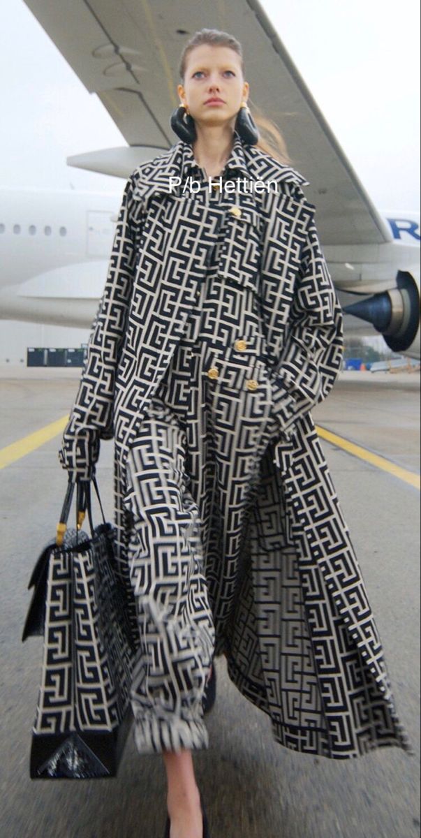a stylish woman wearing a long, black and white geometric print coat strolls through an airport, a large airplane visible in the background. she carries a matching handbag and wears long earrings.  the overall impression is one of high fashion and elegance.