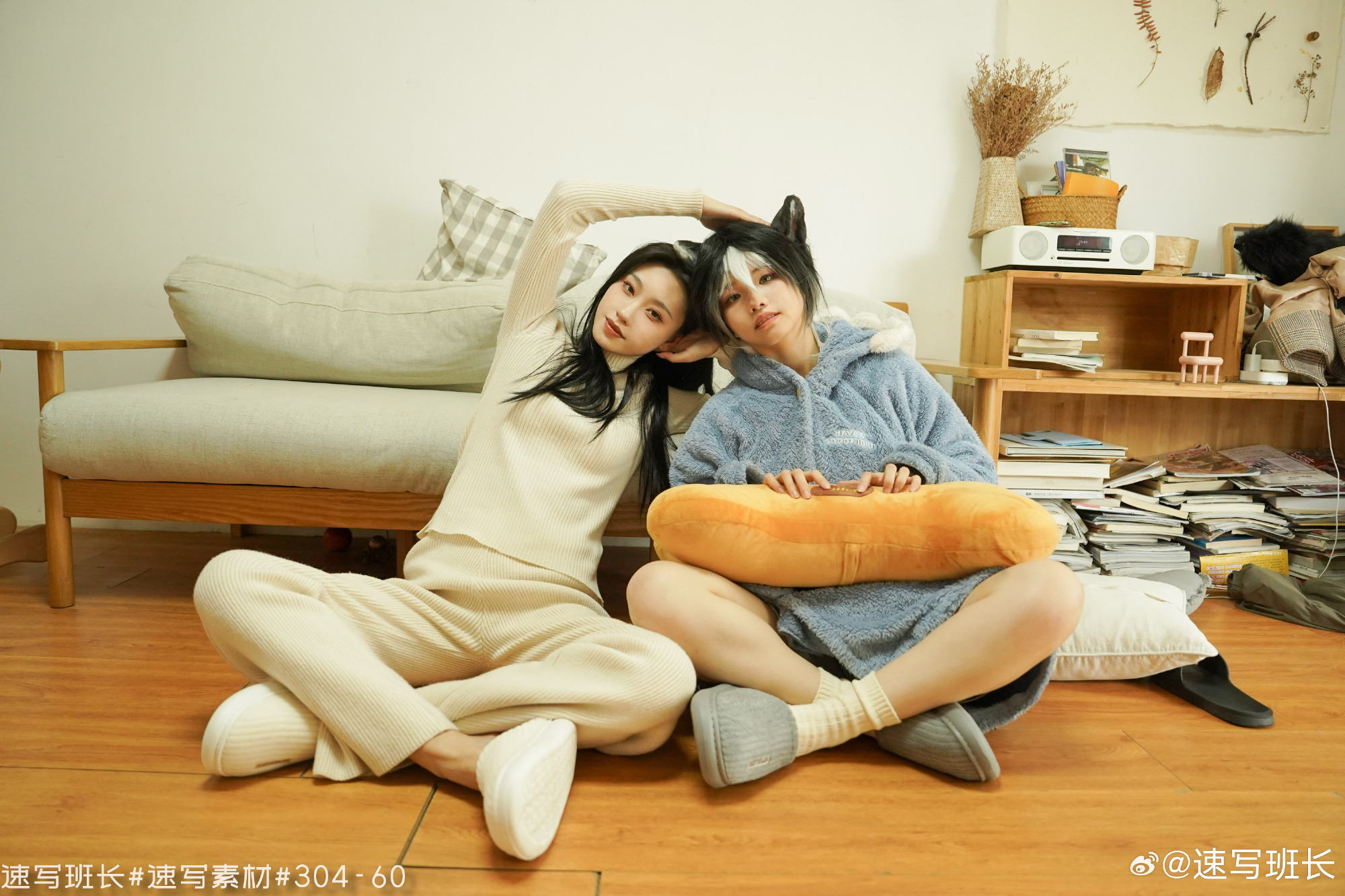 two young women are posing playfully on the floor in matching pajamas. the woman on the right is wearing a hooded onesie with cat ears, while the woman on the left is wearing a matching sweater and pants set. they are both smiling and leaning their heads against each other. the living room is decorated with a comfortable sofa, a bookshelf, and a vintage radio. the lighting is soft and natural, creating a cozy and inviting atmosphere.
