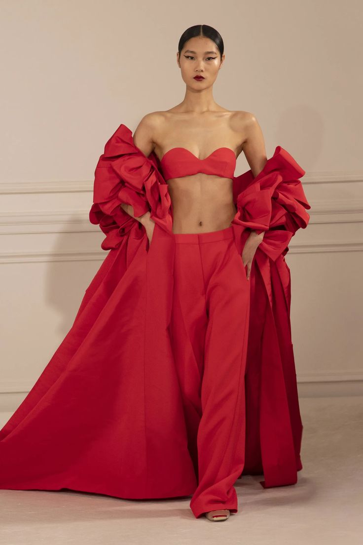 a woman in a red dress with a long, flowing train stands on a runway, striking a pose for a fashion show. the dress has a ruched, voluminous top and wideleg trousers. she is wearing black eyeliner and red lipstick, and her black hair is pulled back from her face. she is wearing high heels. the background is a simple white wall.