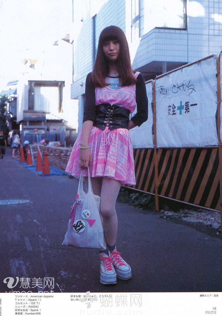 a young woman poses on a city street for a japanese fashion magazine. she is wearing a pink dress over a white tshirt, a black corset belt, white tights, and pink and white platform sneakers. she is carrying a white tote bag. the text on the image is in japanese and includes the name of the magazine and the brands of the clothing she is wearing.