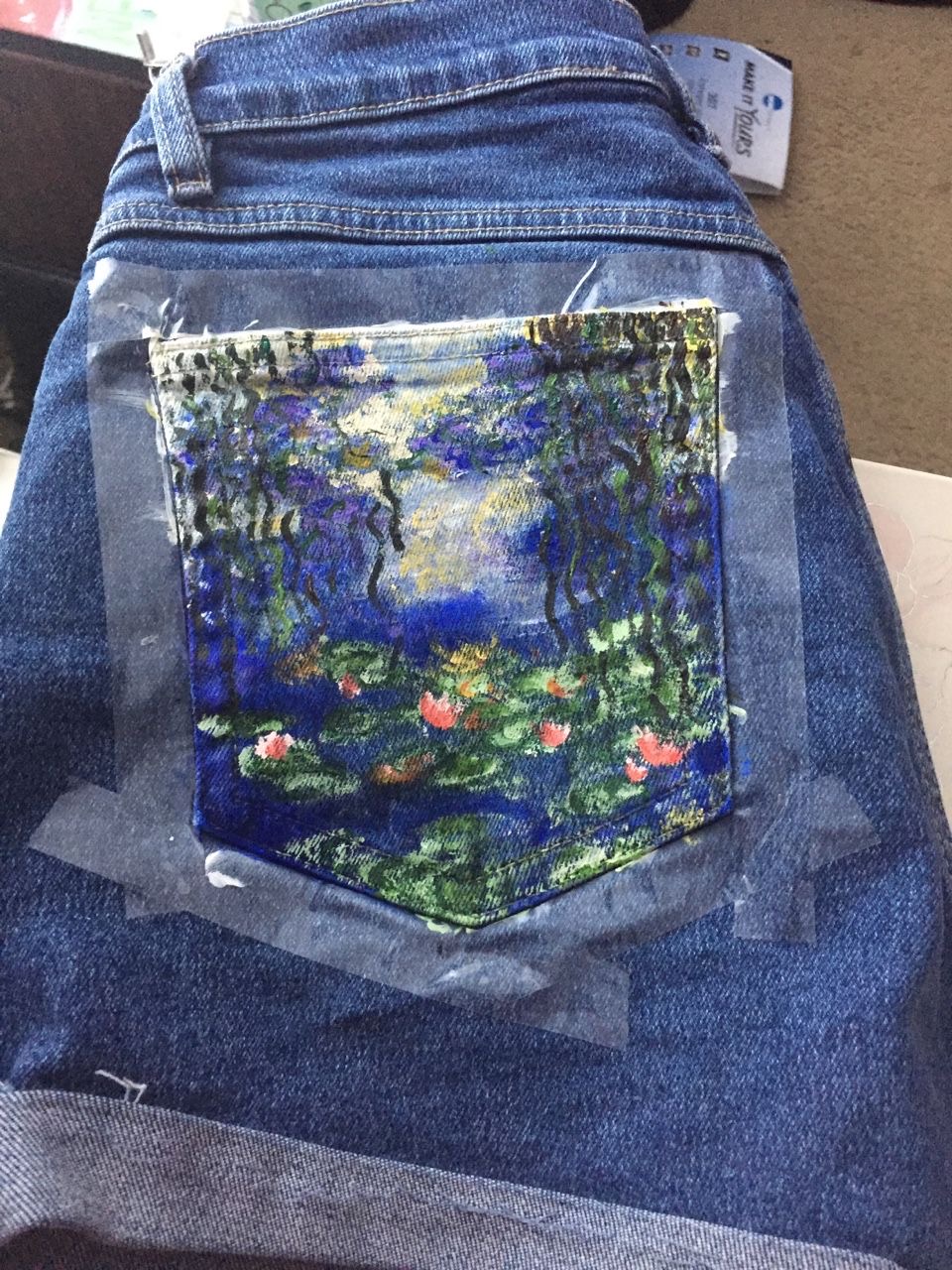 this is an image of a pair of jean shorts laying on a table. the back right pocket of the shorts has been decorated with a handpainted replica of claude monet’s water lilies. the pocket is covered in painter’s tape to protect the rest of the shorts from paint. the pocket is painted predominantly in shades of blue and green with some dabs of pink and yellow.