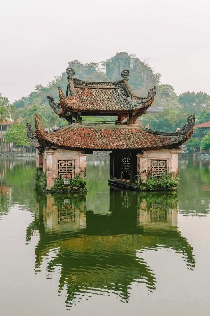 the image shows a beautiful and serene scene of a traditional vietnamese pagoda situated on a lake in ninh binh, vietnam, likely tam coc. the pagoda, with its distinctive east asian architecture and layered roofs, is partially submerged in the calm, green water. its reflection is clearly visible on the surface of the lake, creating a visually appealing symmetry. the background features lush green hills and karst mountains, characteristic of the region. the overall atmosphere is tranquil and peaceful, enhanced by the overcast sky.  the pagoda appears ancient and wellpreserved, suggesting a long history and cultural significance.