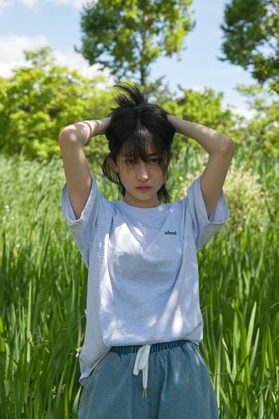 a young woman with short, dark hair is standing in a field of tall green grass. she is wearing a light blue oversized tshirt and denim shorts. the sun is shining brightly, and the woman is looking slightly down. the overall tone of the image is bright and summery.