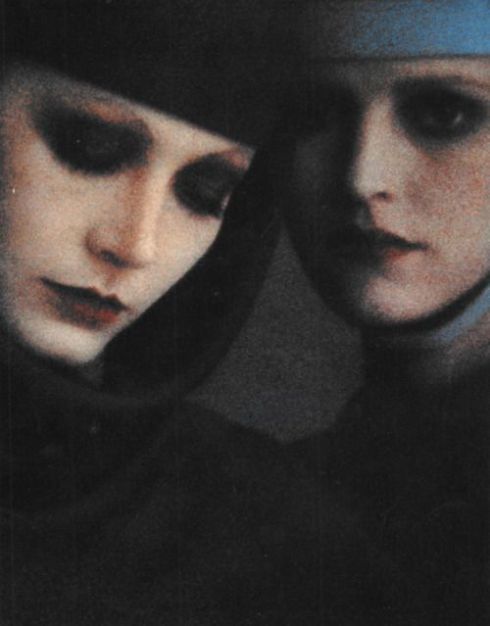 a closeup portrait of two women, side by side, their faces illuminated by a soft, warm light. the woman on the left is wearing a black hat, while the woman on the right wears a blue head covering. their expressions are thoughtful and introspective, their eyes gazing towards the viewer. the overall tone of the image is dark and mysterious, creating a sense of intimacy and connection between the two women.