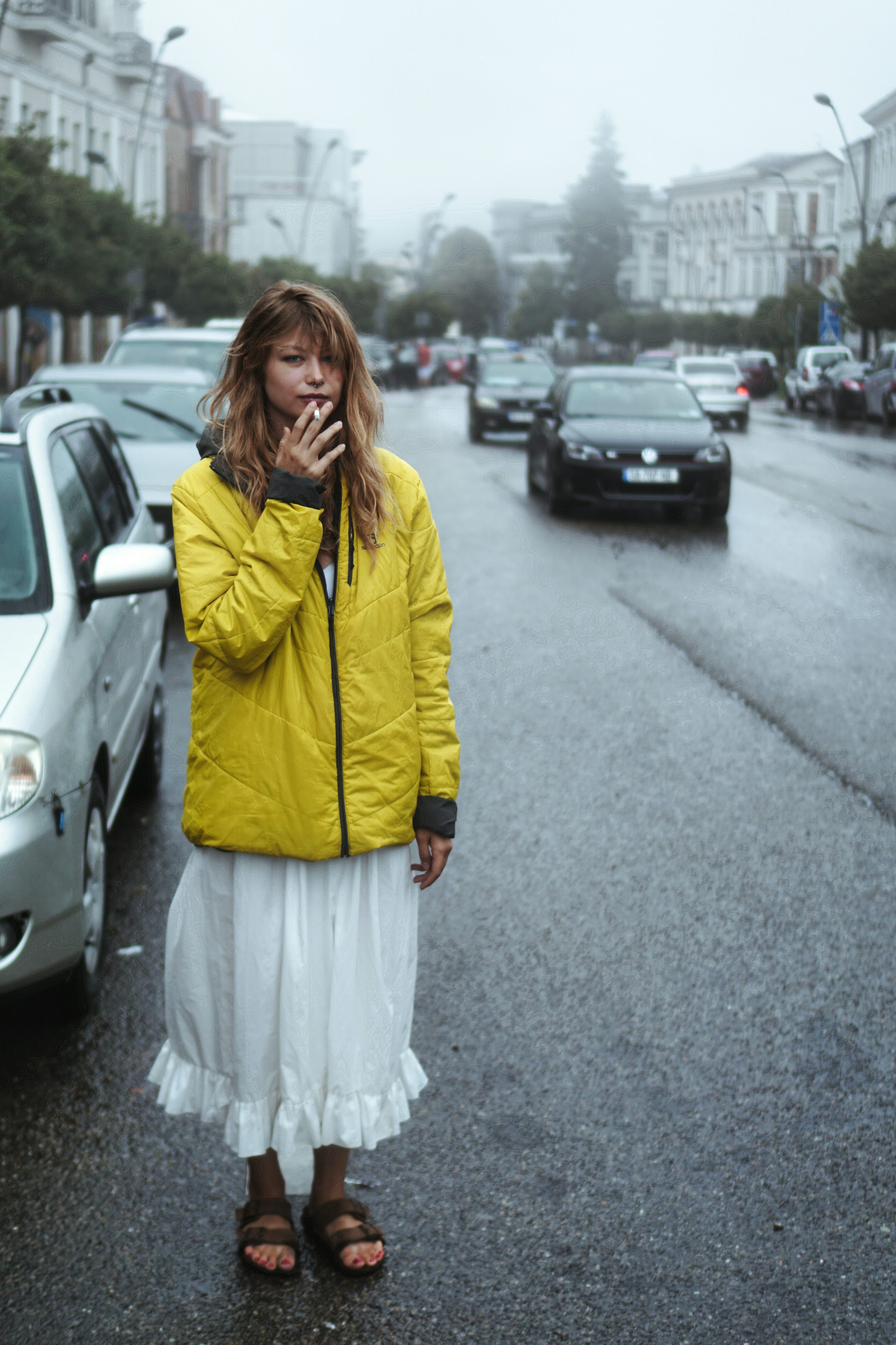 a young woman in a yellow jacket and white skirt stands on a rainy city street, smoking a cigarette. she is looking down, appearing thoughtful or pensive. the background is slightly blurry but shows cars and buildings in a city setting. the overall mood is atmospheric and moody.