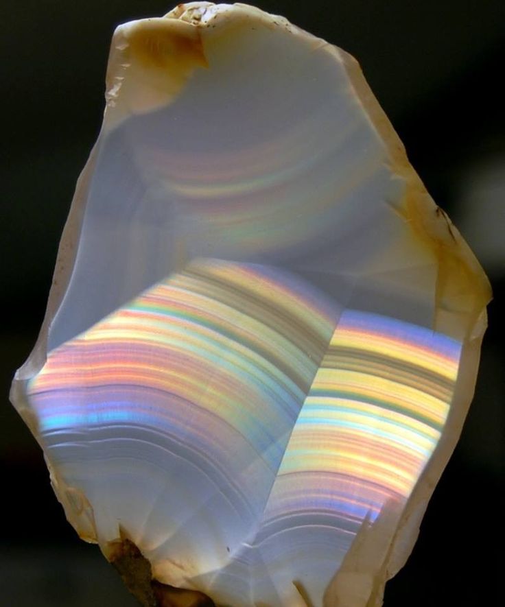 a closeup image of an agate geode with a beautiful rainbow of colors. the agate has a smooth, polished surface, and the iridescent stripes are visible in the center of the stone. the agate is illuminated from the front, highlighting the colorful bands. the geode is resting on a dark background.