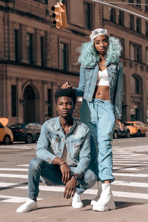 a stylish young african american couple poses in a crosswalk in new york city. the woman has teal hair, is wearing a light blue denim jacket and matching pants, white sneakers and sunglasses. the man is wearing a light blue denim jacket and jeans, and white sneakers. they are both looking at the camera. the background is a city street with buildings and cars. the overall style is urban streetwear.