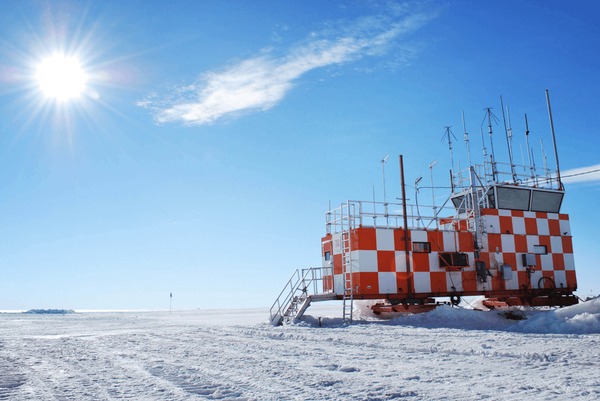 a red and white tower stands on a white snowy landscape with a bright blue sky, sun and clouds. the tower appears to be a research station in antarctica, with a flag on a pole in the distance. it is a very remote and isolated location with a bright and sunny day.  the tower is covered in snow and ice, and the surrounding landscape is also frozen.