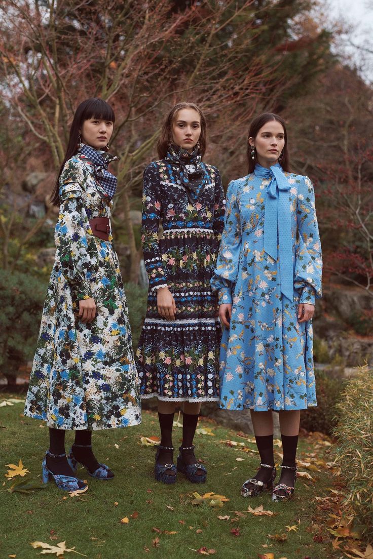 three female models stand in a garden setting, each wearing a different longsleeved floral dress. the model on the left is in a predominantly blue and white floral dress. the center model wears a dark,  multicolored floral maxi dress. the model on the right sports a light blue floral midi dress with a high neck and bow detail.  all three models are wearing dark socks and ankle strap shoes, likely mary jane style.  the overall atmosphere is reminiscent of a fashion photoshoot, set during autumn, in a garden with fallen leaves scattered on the ground. the dresses feature different floral designs, sizes, and cuts suggesting a variety of styles within the same theme.