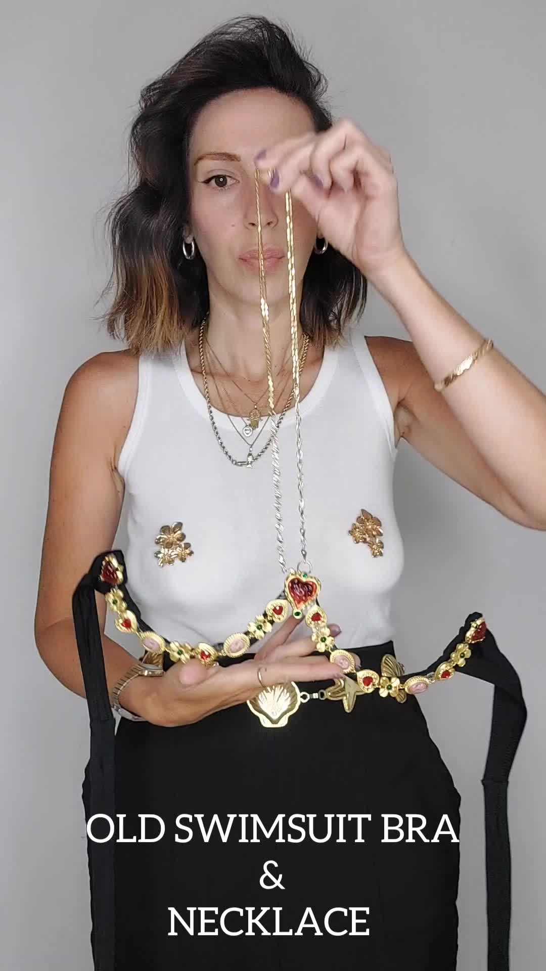 a woman is holding up a gold necklace and a decorated swimsuit bra. she is wearing multiple necklaces and a gold bracelet. she is wearing a white tank top and a black skirt. the text "old swimsuit bra & necklace" is at the bottom of the image.