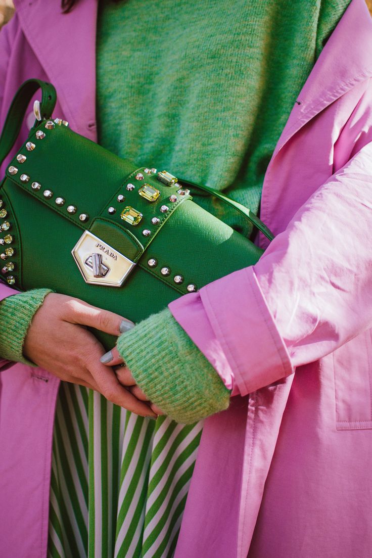 closeup view of a woman holding a green prada handbag adorned with studs and gemstones. she is wearing a pink coat, a green sweater, and a green and white striped skirt. the image showcases a stylish and coordinated outfit, highlighting the details of the handbag and clothing. the focus is on the combination of colors and textures, creating a visually appealing fashion statement.