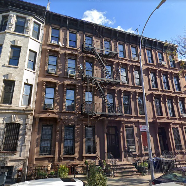 the image shows a row of brownstone apartment buildings on a sunny day. the buildings are three stories tall and have brown brick facades with black fire escapes. there are trees and cars parked along the street in front of the buildings.
