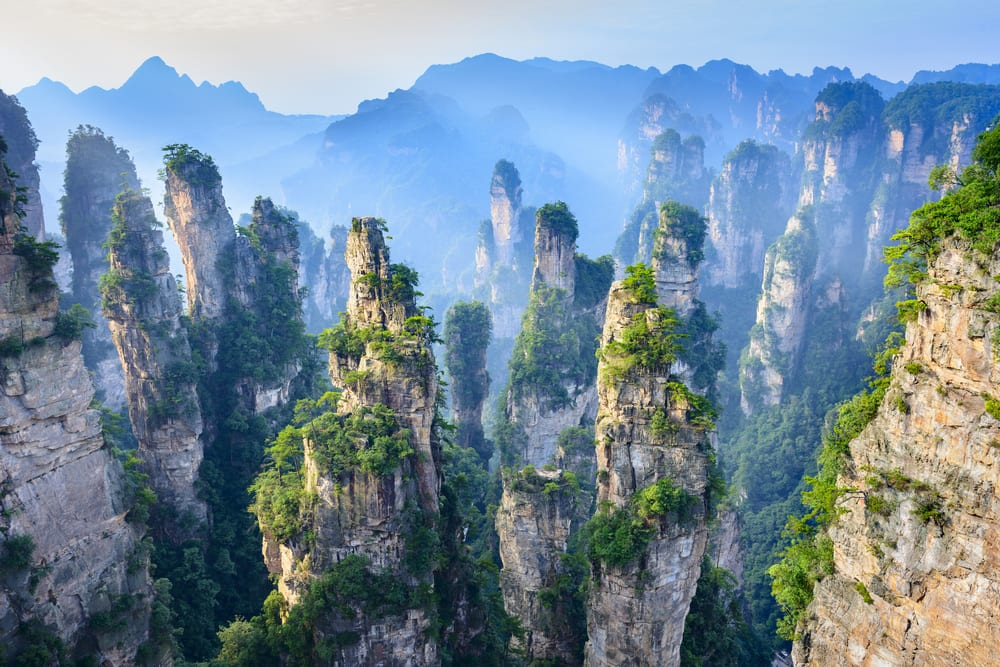 the image showcases the stunning karst landscape of zhangjiajie national forest park in china. towering sandstone pillars rise dramatically from the lush greenery, creating a surreal and majestic scene. mist and fog gently envelop the mountains in the background, adding an ethereal quality to the landscape. the formations are heavily vegetated.