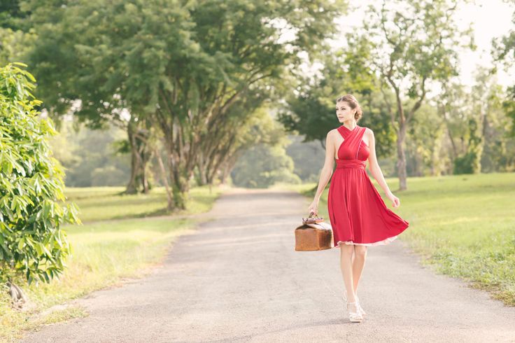 a woman in a red dress is walking down a paved path lined with green grass and trees. she's carrying a brown suitcase and looking off to the right side of the frame. the sun is shining and the overall lighting is warm and inviting.
