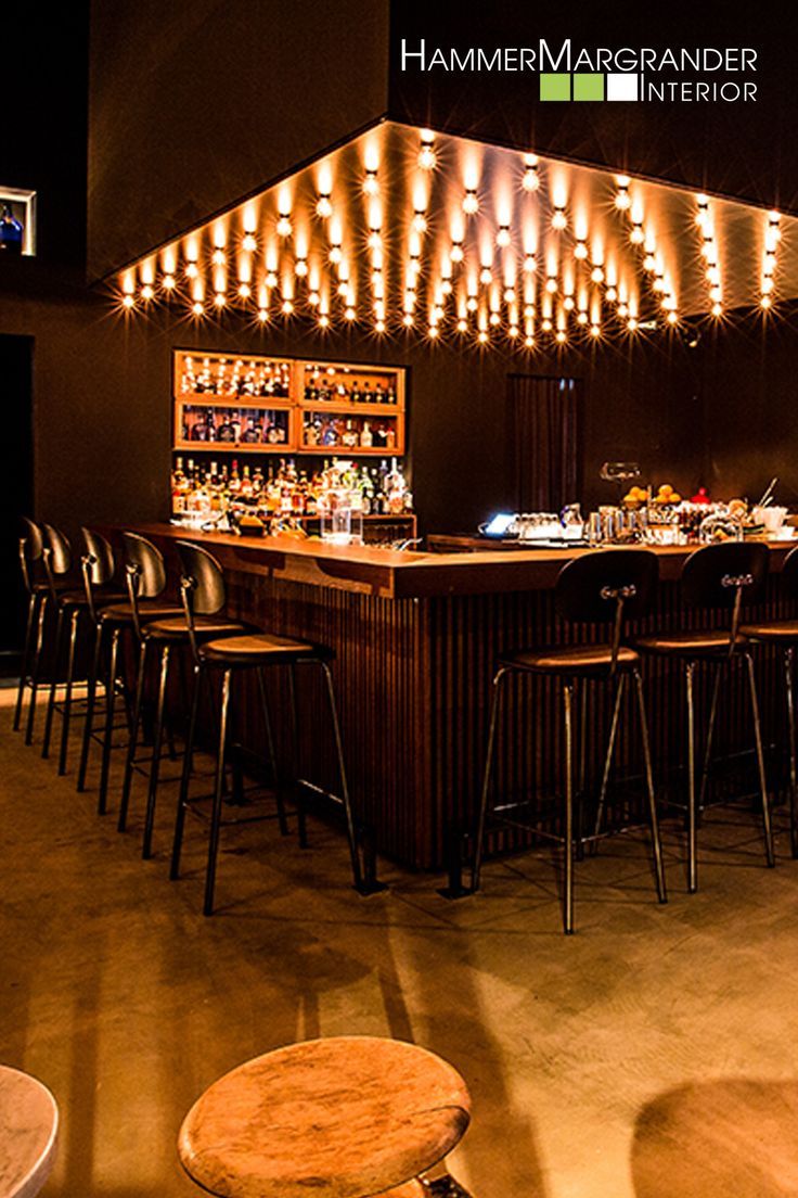 a dimly lit bar with a long wooden counter and black metal stools. warm lights hang from the ceiling, illuminating the bottles and glasses behind the bar. the overall aesthetic is modern and industrial with a warm and inviting atmosphere.