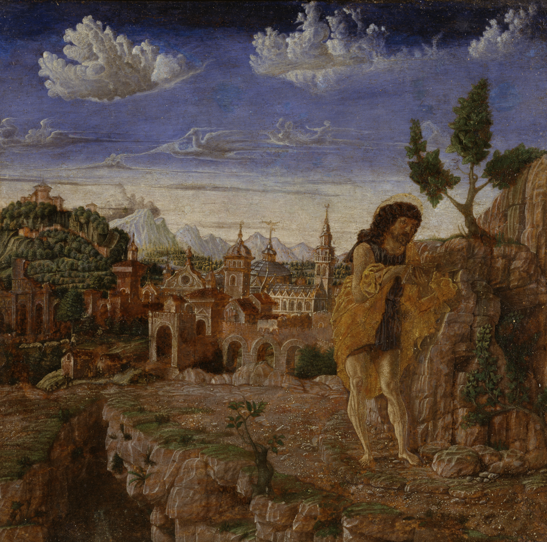 this is an old master painting depicting st. john the baptist in a wilderness setting with a city in the background. the painting shows a bearded man with a halo around his head standing near a rock formation, wearing a simple garment. he appears to be looking down or concentrating on something in his hands. behind him, there is a landscape with a city featuring various buildings, including towers and arched structures, set against a backdrop of mountains and a cloudy sky. the terrain around the figure is rugged, with a cliff and sparse vegetation.