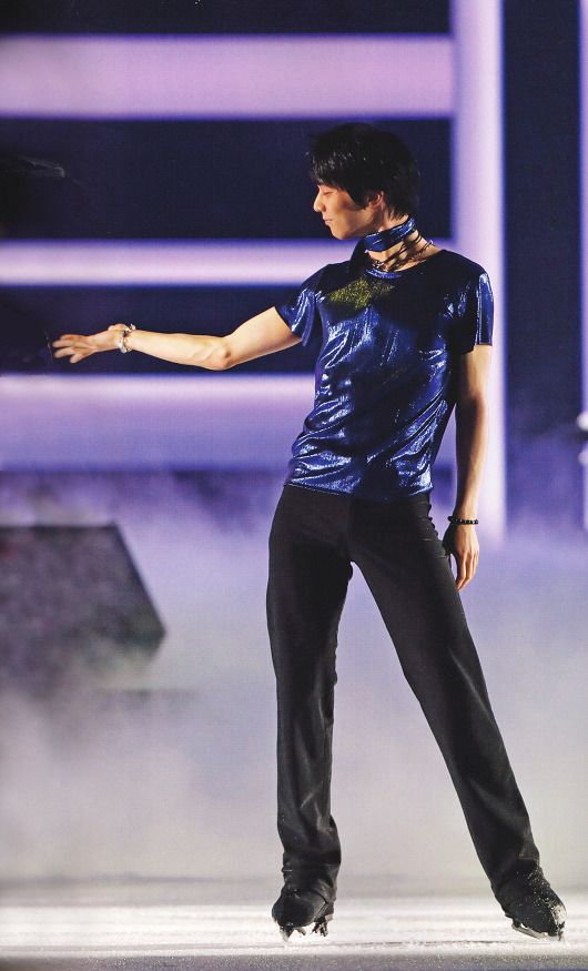 a male figure skater wearing black pants and a blue shirt skates on a sheet of ice during a competition. the skater is poised with a determined expression, and his skates are clearly visible. the background features a blurred image of the arena, including the stands and the ice surface. the lighting is bright and dramatic, highlighting the skater's form and movement.