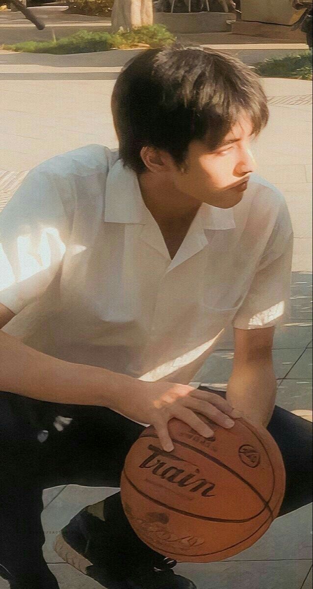a young man, bathed in warm sunlight, holds a basketball as he crouches on a sidewalk. his white shirt and dark pants contrast with the light pavement and green foliage in the background. the image evokes a sense of leisure and athleticism.