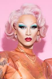 the image is a closeup portrait of a drag queen. she has a voluminous, pale pink hairstyle reminiscent of a vintage hollywood glamour style.  her makeup is bold and precise, featuring dramatic teal eyeshadow, long false eyelashes, and a vibrant red lipstick. she's wearing a peachcolored translucent outfit with shoulder pads, and a gold necklace is visible around her neck. the background is a solid, pale pink.  part of a tattoo is visible on her arm. the overall aesthetic is glamorous and slightly retro.
