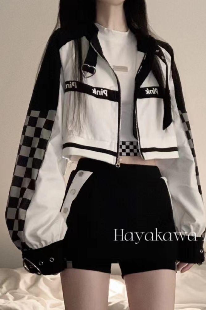 the image shows a model wearing a stylish black and white cropped jacket with a checkered pattern on the sleeves. the jacket features pink lettering that says "pink" and a small heartshaped detail.  she is also wearing highwaisted black shorts. the overall style is streetwearinspired and trendy. the model's hair is dark and straight.  the background is a muted beige.