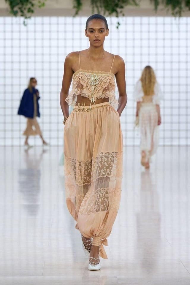 a woman models a fashion outfit on a runway. the outfit consists of a crop top with lace trim, a matching highwaisted skirt with lace inserts and a thin belt. the model is wearing white shoes. in the background, there are other models and runway staff. the background is a white studio with greenery hanging above.